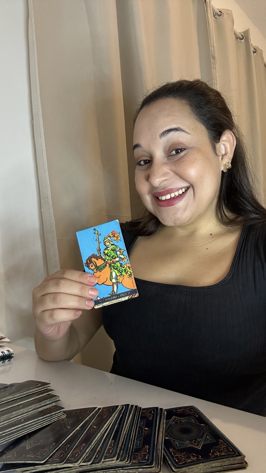 Pamela Abreu sorrindo, com uma carta de Tarot e seu baralho sobre a mesa